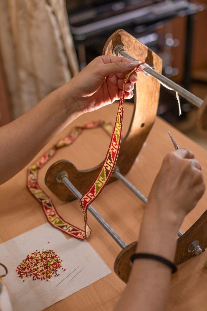 A close-up of hands crafting detailed embroidered trim using a traditional spindle tool, with red and gold thread, highlighting the craft of luxury at Style Haven.