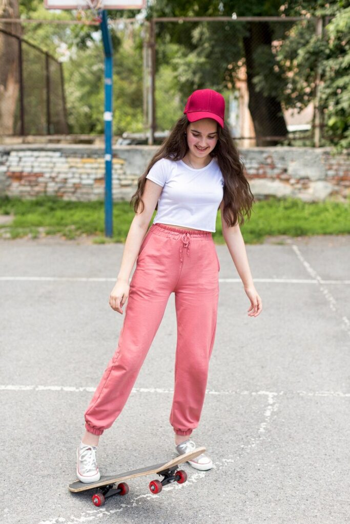 "Smiling young woman in pink joggers and a red cap balancing on a skateboard in an urban park setting."