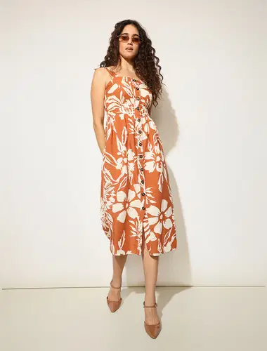 Stylish woman wearing an orange floral midi dress with neutral heels, posing against a plain studio background."